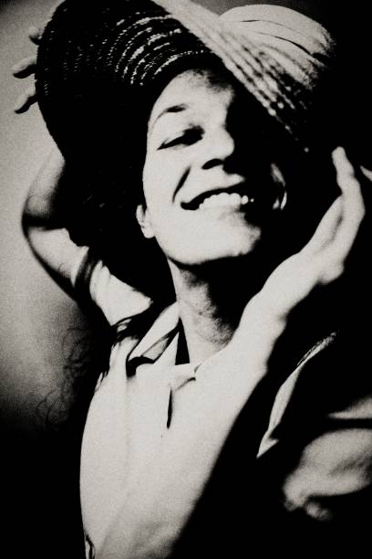 Black and white photo of a woman putting on a straw hat. She smiles at the viewer.