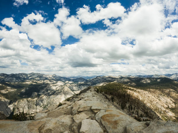 A breathtaking panoramic view from the summit of Half Dome in Yosemite National Park, capturing the vast expanse of rugged mountain ranges under a sky filled with dramatic clouds.