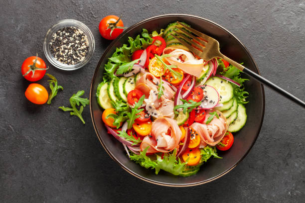 istock Salad with fresh vegetables and jamon in a black bowl on the table. Top view 2171518465