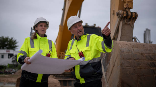 istock Construction workers are standing in front of heavy machinery (an excavator) and examining a large blueprint or construction plandiscussing the project details concept. 2171518706