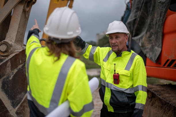 istock Construction workers are standing in front of heavy machinery (an excavator) and examining a large blueprint or construction plandiscussing the project details concept. 2171518747