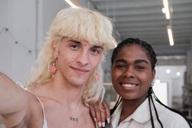 waist up shot of a professionals freelance looking at the camera and smiling. non binary caucasian person and a Latino black female standing  indoors on a conceptual handcraft studio
