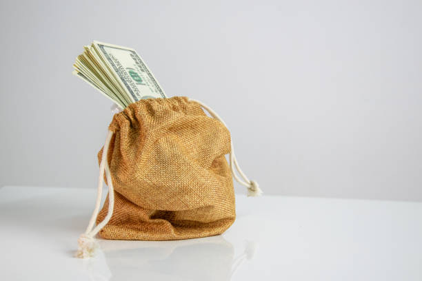 A burlap sack with a drawstring sits on a white surface with a stack of US dollar bills spilling out from the opening.