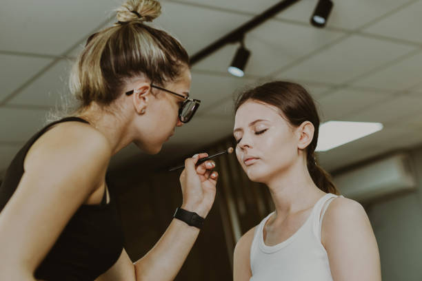 One young handsome Caucasian makeup artist applies highlighter with a brush to the right cheekbone of the face of a girl sitting in a chair early in the morning in a beauty salon, close-up view from below. Step by step.
