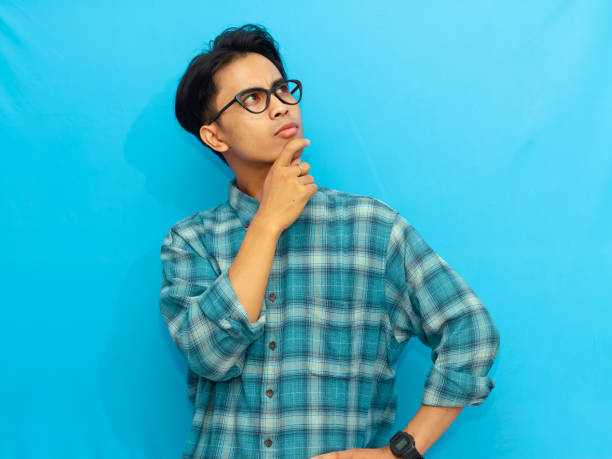 istock Asian man in a blue casual shirt posing with a serious thinking expression, looking up with eyes focused, set against a blue background. 2171577876