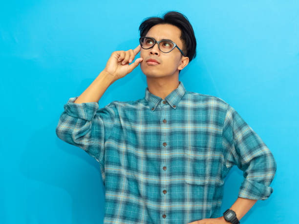 istock Asian man in a blue casual shirt posing with a serious thinking expression, looking up with eyes focused, set against a blue background. 2171577896