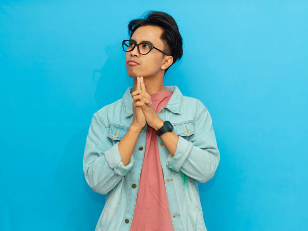 istock Asian man wearing a denim jacket posing thoughtfully eyes looking up isolated on a blue background. 2171603356