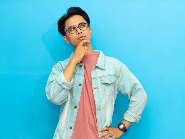 istock Young Asian man in casual clothing posing with a serious expression thinking while looking up isolated on a blue background. Perfect for advertising or promotional content. 2171604309