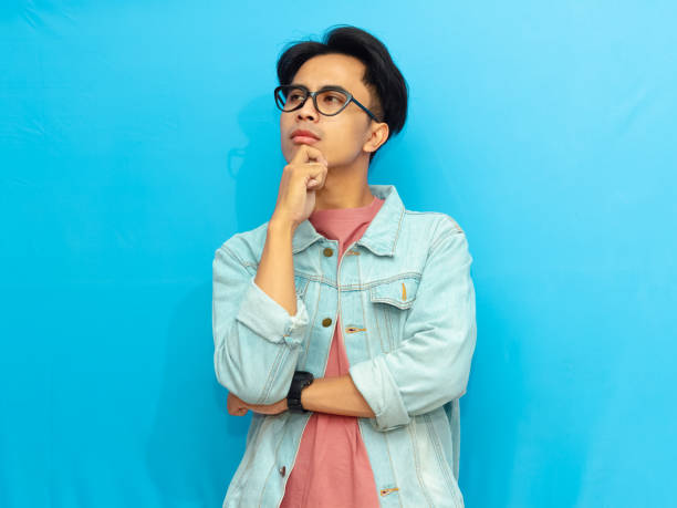 istock Young Asian man in casual clothing posing with a serious expression thinking while looking up isolated on a blue background. Perfect for advertising or promotional content. 2171604311