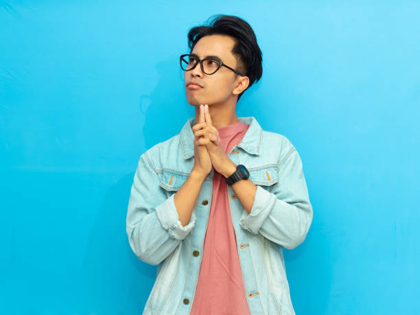 Young Asian man in casual clothing posing with a serious expression thinking while looking up isolated on a blue background. Perfect for advertising or promotional content.