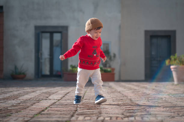 One and half year old multiracial baby boy in a vibrant red sweater, wearing a brown beanie, delights in a playful run across a sunlit cobblestone courtyard with potted greenery and a simple building facade. Christmas spirit outfit