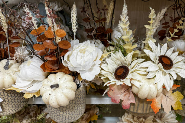 a decorative autumn-themed floral arrangement featuring white pumpkins, artificial white flowers, wheat stalks, and dried leaves