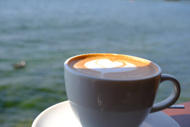 Close-up view of cafe latte in white cup by the sea on a sunny summer day. There is the sea in the background. Weekend coffee chat with friends.
