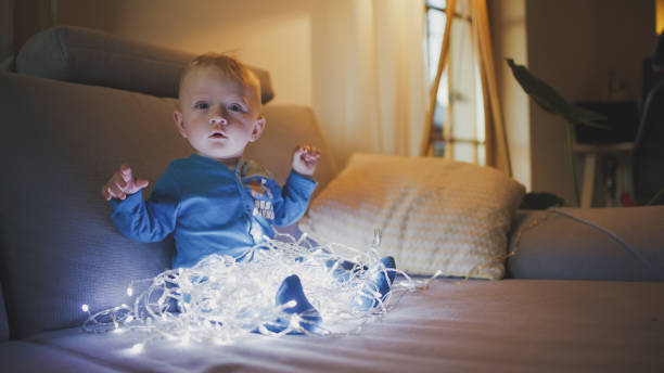 A young Caucasian boy delightfully engages with twinkling Christmas lights, sitting comfortably at home in casual attire, surrounded by soft furnishings.