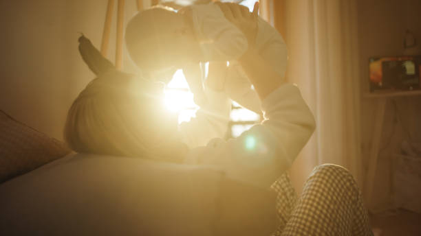 A caring Caucasian woman lifts her baby boy in the air, their bond evident in a well-lit, cozy indoor setting. Both are dressed in comfortable home attire, embodying a serene, nurturing interaction.