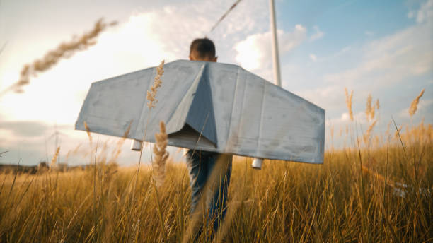A young Caucasian boy dashes through a field, playful joy on his face as he holds a toy airplane, with towering wind turbines under a vast sky in the backdrop.
