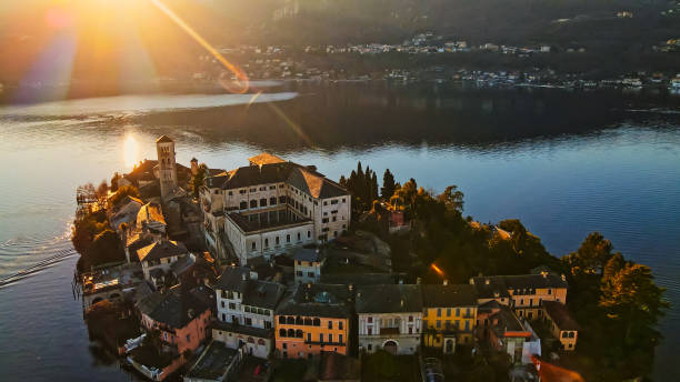 Aerial view highlights Island San Giulio bathed in golden sunrise light with surrounding calm waters reflecting the warm hues, emphasizing the peaceful ambiance of this iconic location.
