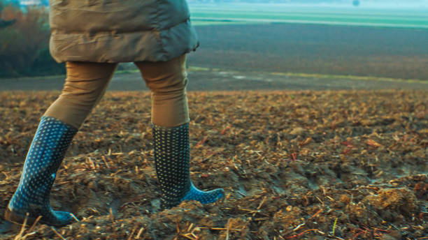 A mid adult Caucasian woman in boots treads carefully across a plowed agricultural field, exemplifying rural life and agriculture profession.