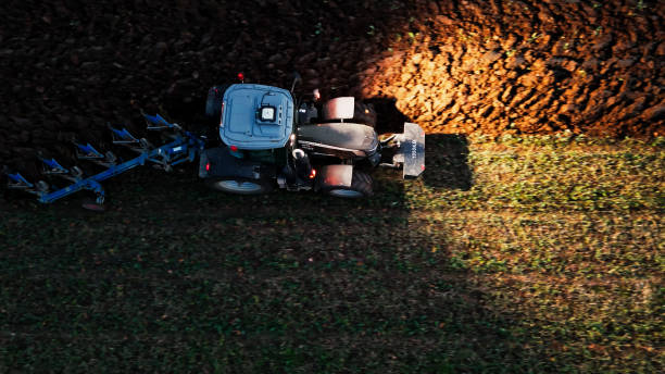 Overhead view of a diligent farmer maneuvering a modern tractor at night, its lights illuminating the rich soil on an expansive agricultural field.