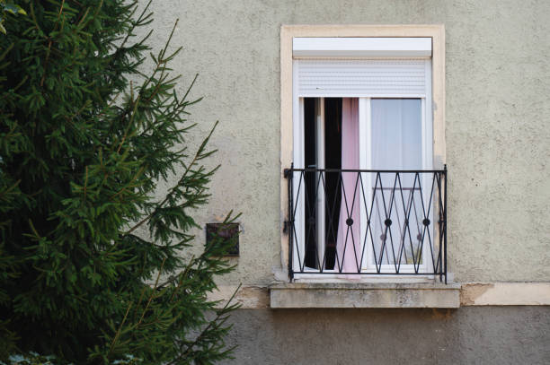 An old apartment building facade with a small balcony featuring a hanging cloth. The walls show signs of wear, with cracked plaster and vines growing up the side. Lace curtains cover the windows, adding a nostalgic touch to the weathered exterior.