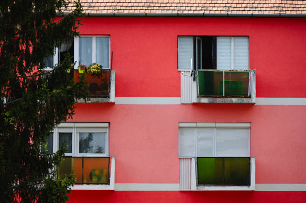 A vibrant pink apartment building featuring small balconies with green-tinted glass railings. Some balconies are adorned with plants, while others remain simple. The bold contrast between the pink facade and green balconies creates a striking visual appeal, enhanced by a nearby tall tree.