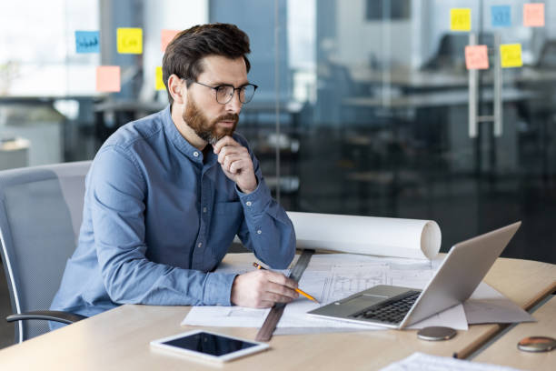 istock Serious and thoughtful young man architect, engineer sits in the office with projects and looks at the laptop screen 2171671269
