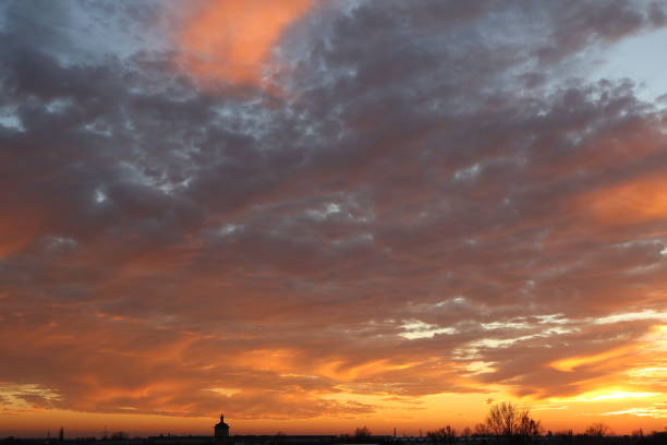 A serene sunset graces the sky, painting it with vibrant hues of orange, pink, and purple. Wispy clouds add texture to the scene, creating a mesmerizing pattern. The silhouette of trees and small structures is visible on the horizon, adding depth and balance to this captivating natural spectacle.