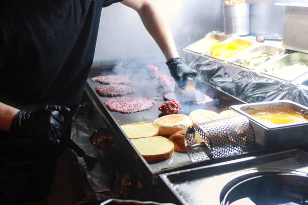 Chef preparing smash burgers on a hot griddle, pressing meat patties and toasting buns Chef preparing smash burgers on a hot griddle, pressing meat patties and toasting buns, capturing the fast-paced cooking process in a busy commercial kitchen. spanish hamburger stock pictures, royalty-free photos & images