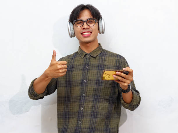 istock Young happy Asian man playing mobile games or watching digital video streams on his smartphone, giving a thumbs-up and OK sign, isolated on a white background, perfect for advertising. 2171702644