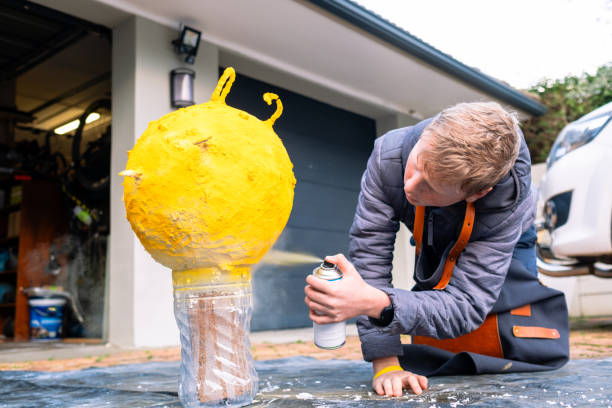 A young boy uses a can of yellow spray paint to color his model of the sun for his school science project. The sun has been made from layers of paper mache pasted together with glue