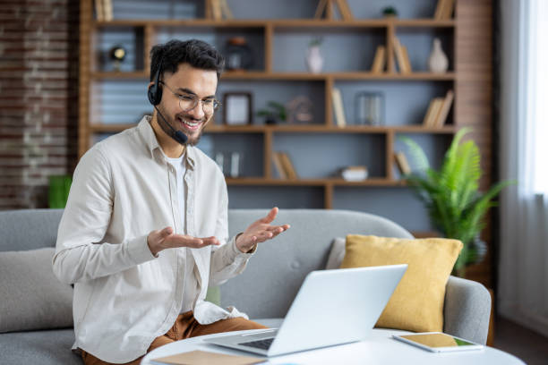 istock Smiling young Indian man sitting on sofa at home wearing headset, talking on video call, gesturing with hands 2171721682