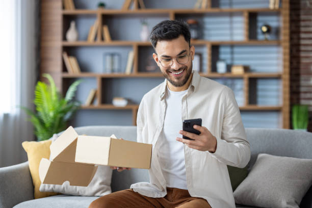 istock Happy man unboxing parcel and using smartphone in living room 2171721804