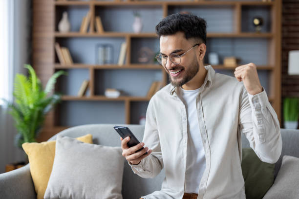 istock Smiling man celebrating success while using smartphone on couch at home 2171721822