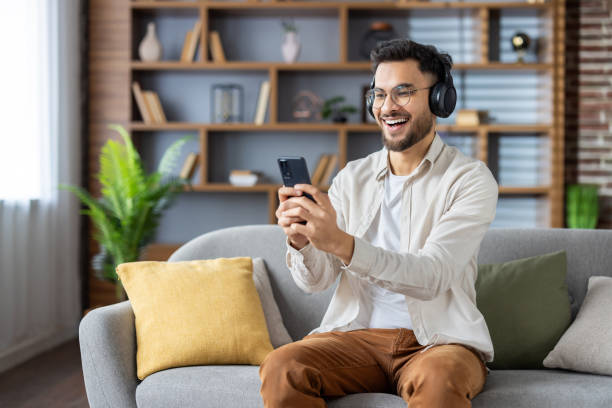 istock Smiling man enjoying music on headphones while using smartphone at home, relaxing on sofa. Tech-savvy individual immersed in digital entertainment. 2171735261