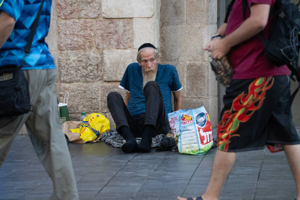 Jerusalem, Israel - July 18th, 2024: A fingerless beggar sitting on the sidewalk on a Jerusalem street, as people pass him by.