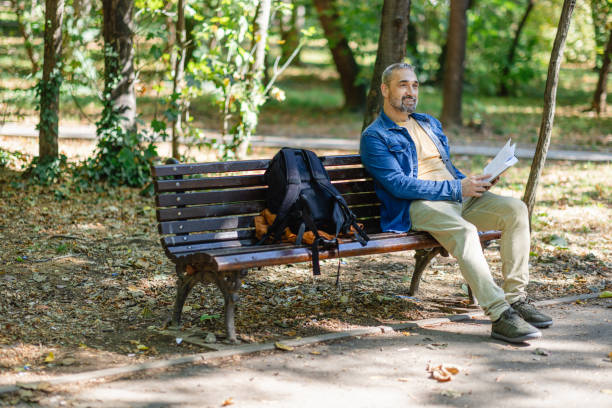 A man sits comfortably on a wooden bench in the park, engrossed in reading papers, with his backpack resting beside amid a backdrop of trees.