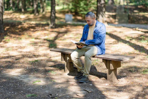 A man dressed in a denim jacket and beige pants sits alone on a wooden bench, engrossed in reading a book in the serene and leafy environment of a public park.