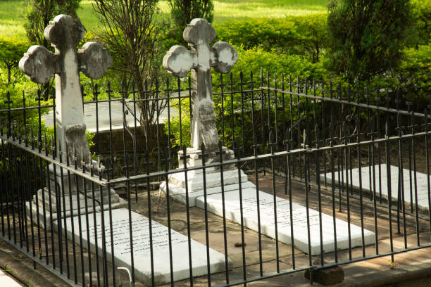 A weathered stone cross is prominently displayed in a cemetery, surrounded by headstones and trees, under a gray and cloudy sky in the late afternoon light.