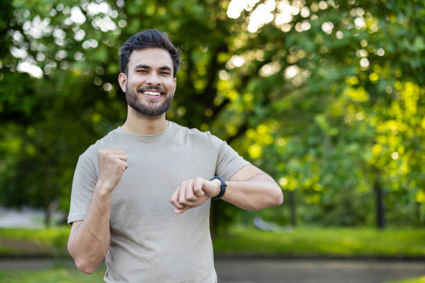 istock Energetic young man in park checking fitness tracker after exercise session, smiling confidently in casual attire beneath vibrant green trees 2171765251