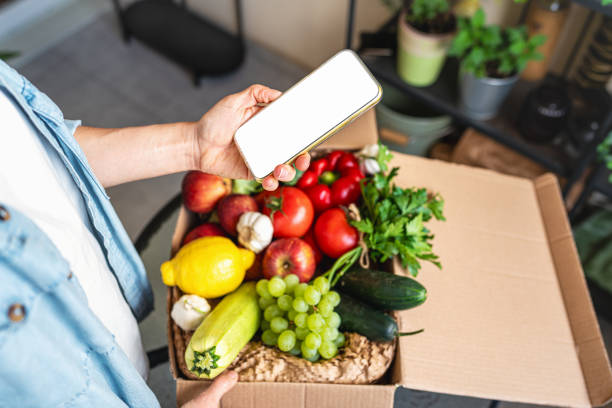 istock Mockup mobile app ordering farm grown vegetables. Mobile phone with blank screen in female hand above the box with fresh and ripe organic vegetables. 2171768009