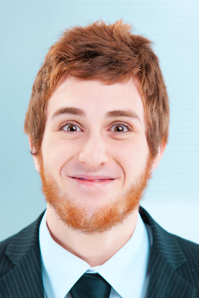 Young businessman with a big red beard smiles awkwardly, exuding positivity and joy in the office. His goofy expression adds humor to the workplace