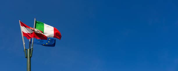 Italian, Austrian, and European Union flags flutter together on a flagpole against a clear blue sky, symbolizing unity and cooperation