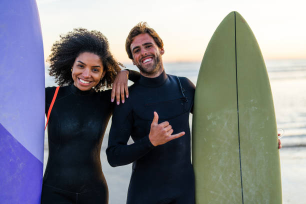 A smiling black woman and a white man in wetsuits stand with surfboards, exuding a relaxed beachside vibe as the sun sets.