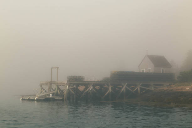 A small wooden pier in Maine extends into the calm water, partially obscured by a thick layer of fog. A quaint house sits at the end of the pier, adding to the serene and mysterious atmosphere.