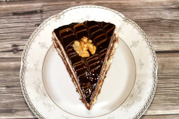 chocolate triangular spongy cake piece filled and topped with walnut, layers of chocolate paste cream and pieces, with layers of whipped cream, served in a plate and isolated on wooden background