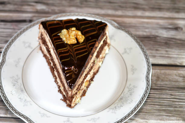 chocolate triangular spongy cake piece filled and topped with walnut, layers of chocolate paste cream and pieces, with layers of whipped cream, served in a plate and isolated on wooden background