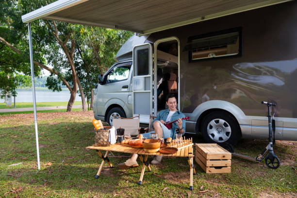 Young Asian man enjoy playing ukulele while sitting on foldable chair during breakfast time beside the camper trailer.