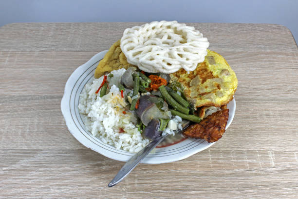 Nasi sayur lodeh, a typical Indonesian lunch or breakfast, rice with vegetables cooked in coconut milk sauce with side dishes of omelette, tofu, tempeh, crackers, and chili sauce, served on a white ceramic plate on a wooden patterned table, eaten while still hot or warm