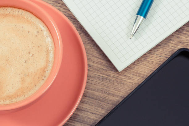 White cup with tea on a saucer with cookies next to an open blank notebook and a pen on a dark wooden table