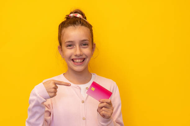 In front of yellow wall cheerful girl holding credit card and pointing with finger on it